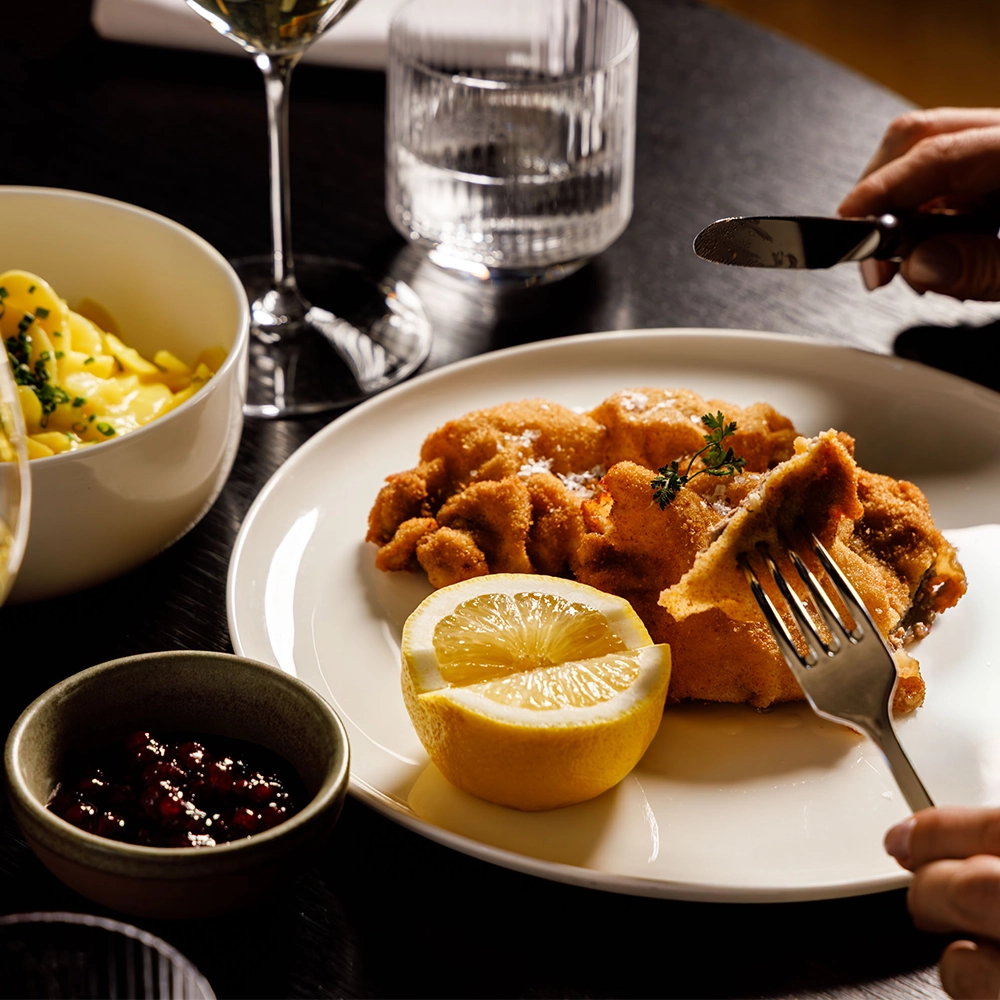 Wiener Schnitzel mit Zitronenspalte und Preiselbeeren, serviert mit Kartoffelsalat und einem Glas Weißwein in stilvollem Restaurantambiente.